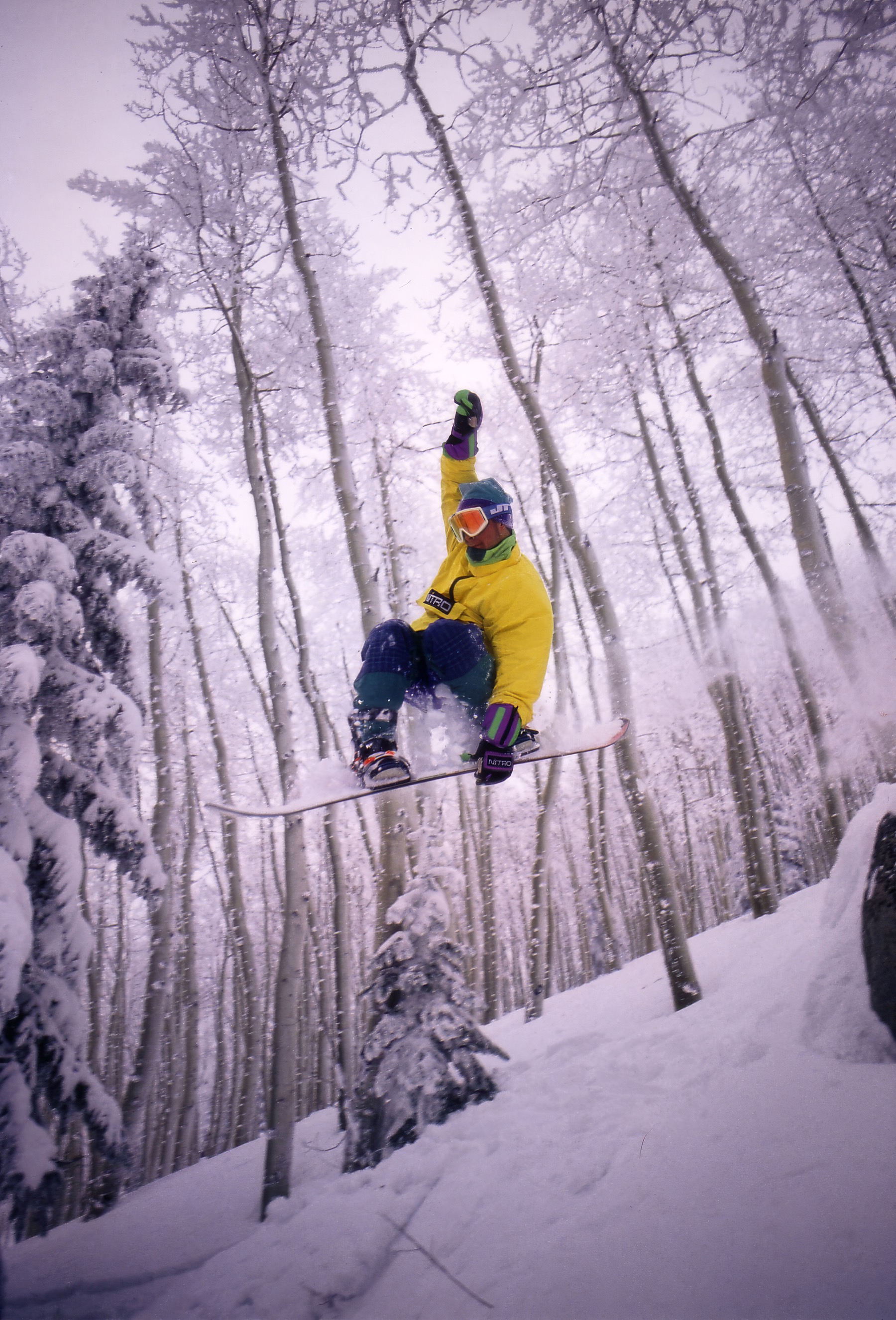 Snowboarder in bright yellow gear jumping through a snowy aspen forest.